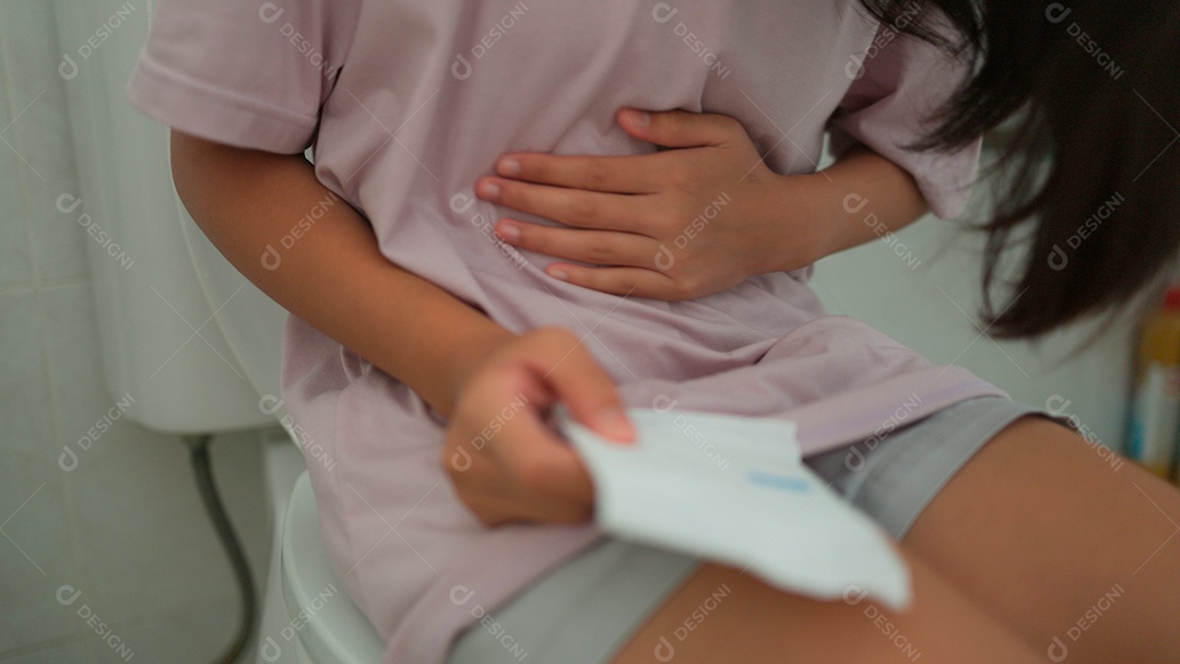 Uma menina asiática doente sentada no vaso sanitário do banheiro e segurando seu estômago doloroso durante o período menstrual