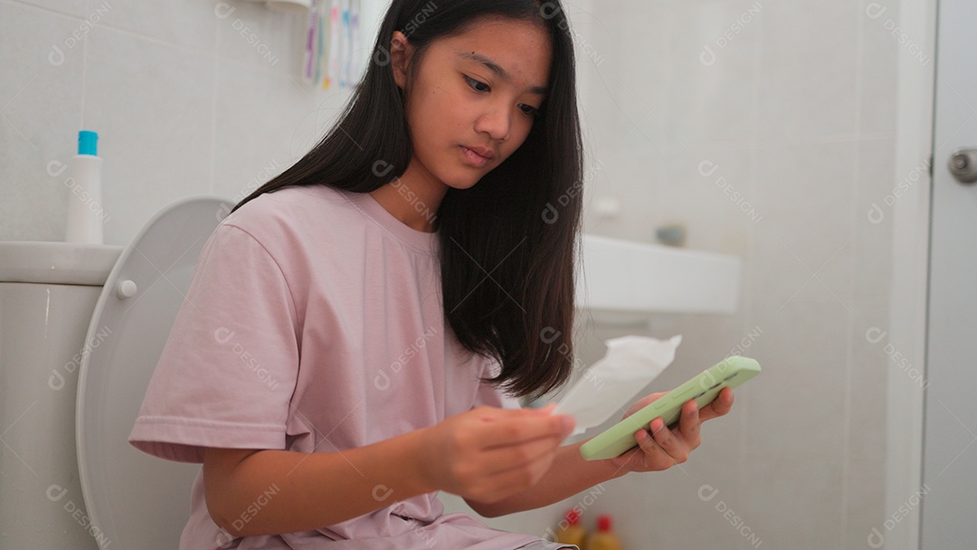 Uma menina asiática doente sentada no vaso sanitário do banheiro e segurando seu smartphone no período mestrual