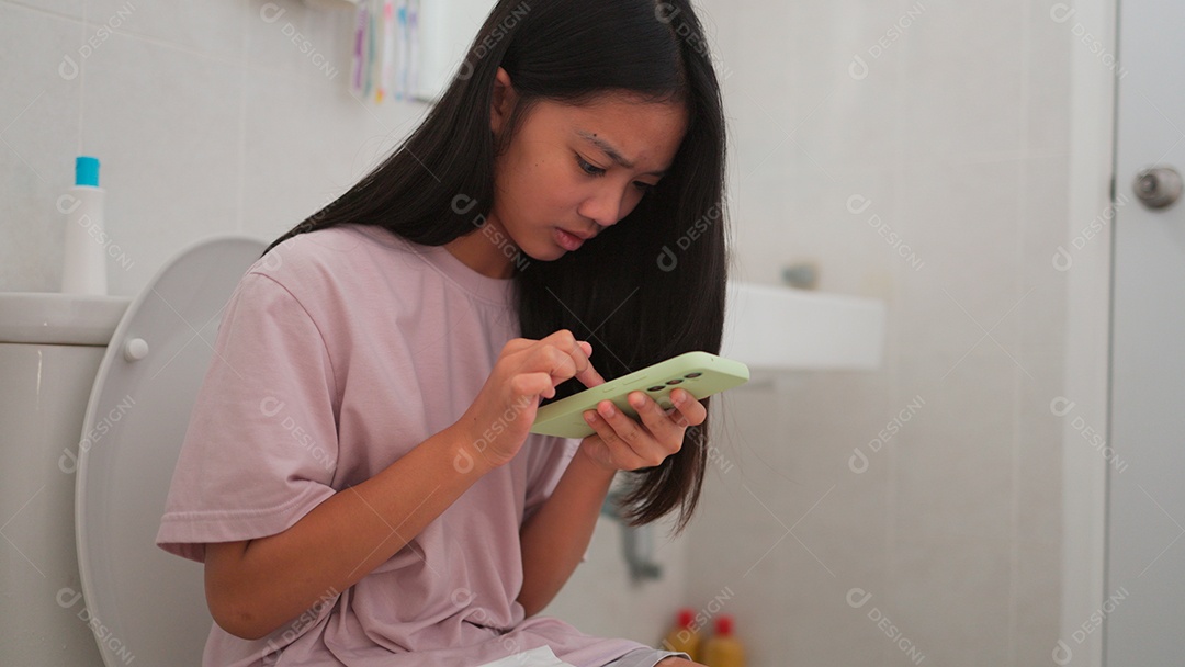 Uma menina asiática doente sentada no vaso sanitário do banheiro e segurando seu smartphone no período mestrual