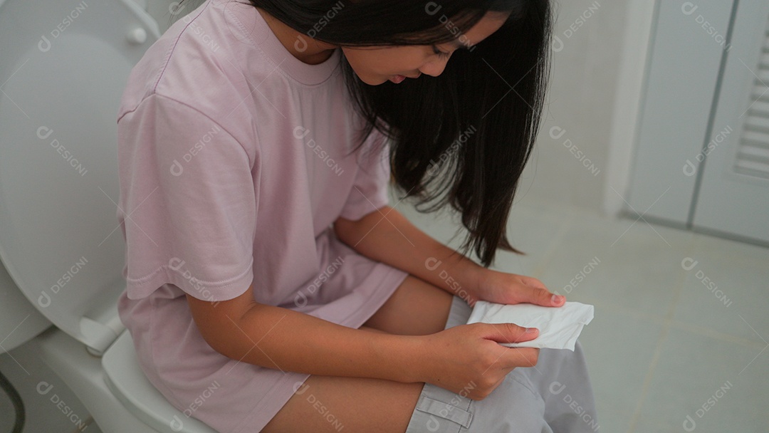 Uma menina asiática doente sentada no vaso sanitário do banheiro e segurando seu absorvente no período mestrual