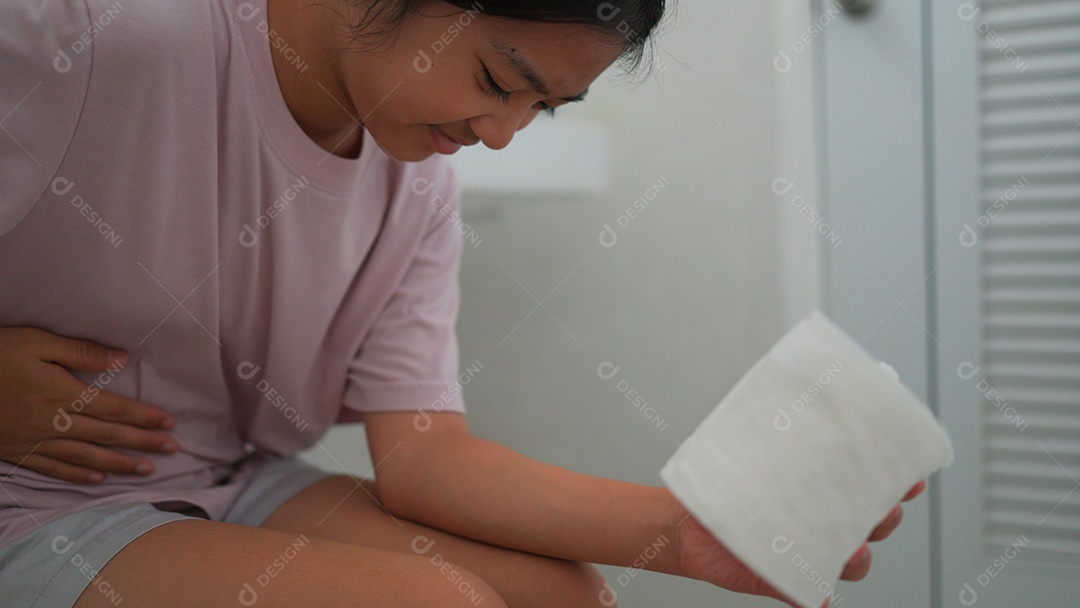 Linda menina asiática sentada em vaso sanitário segurando papel higiênico com dor de barriga