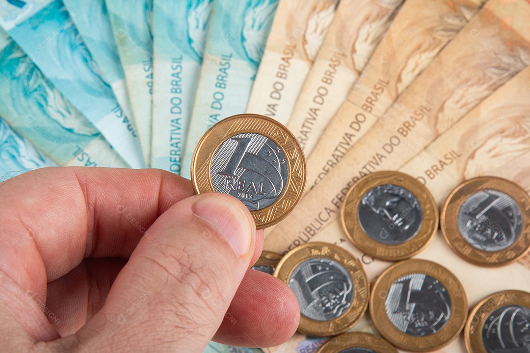 Pessoa segurando moeda de um real brasileiro contra fundo desfocado de notas