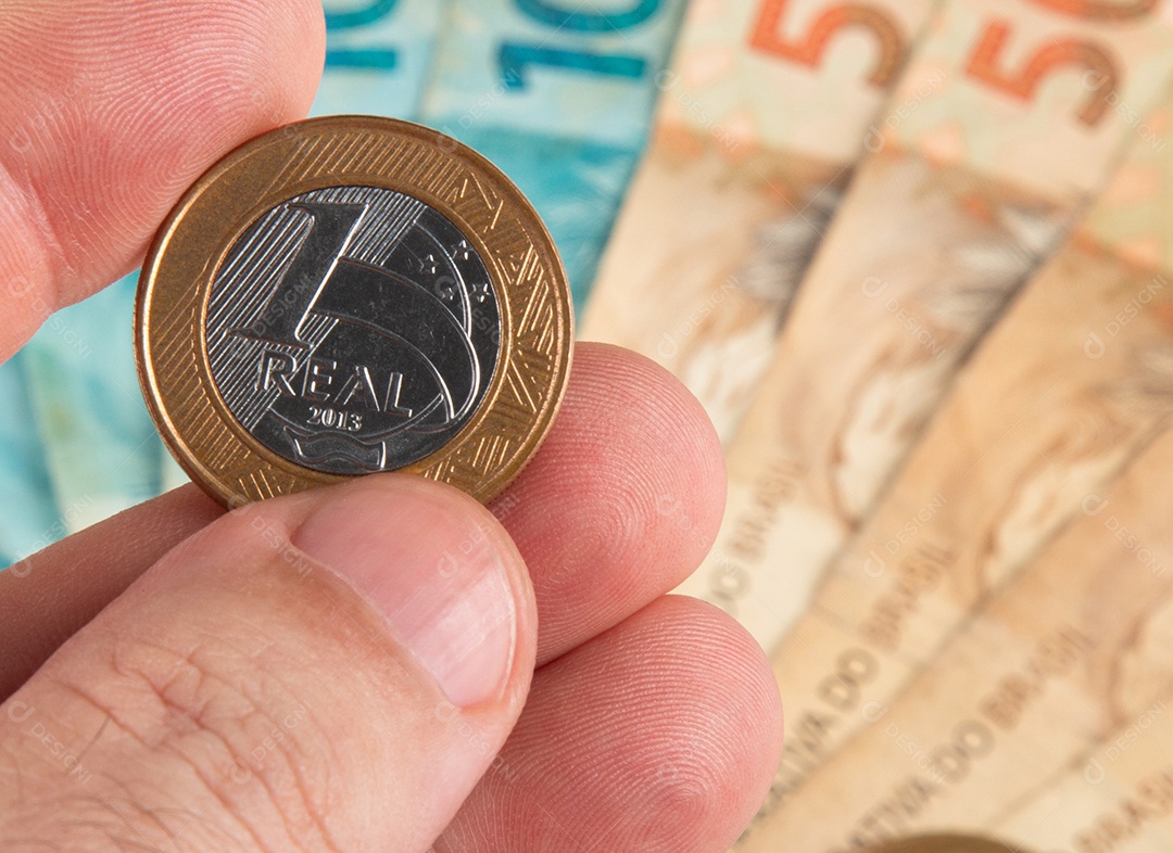 Pessoa segurando moeda de um real brasileiro contra fundo desfocado de notas