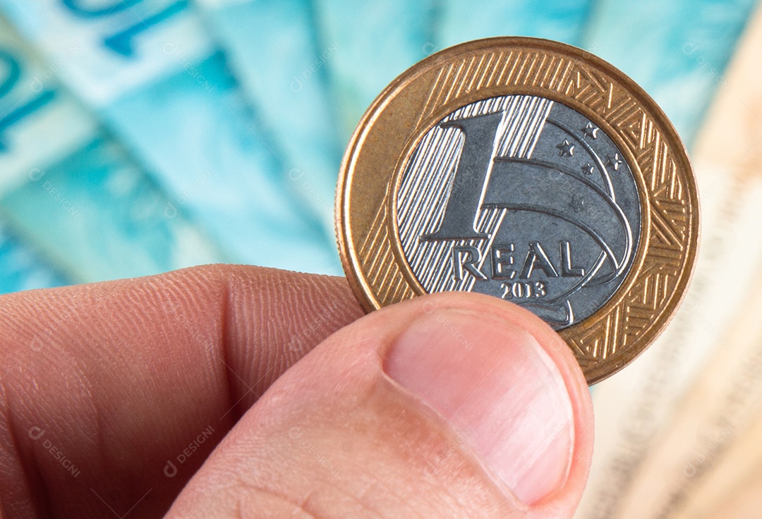 Pessoa segurando moeda de um real brasileiro contra fundo desfocado de notas