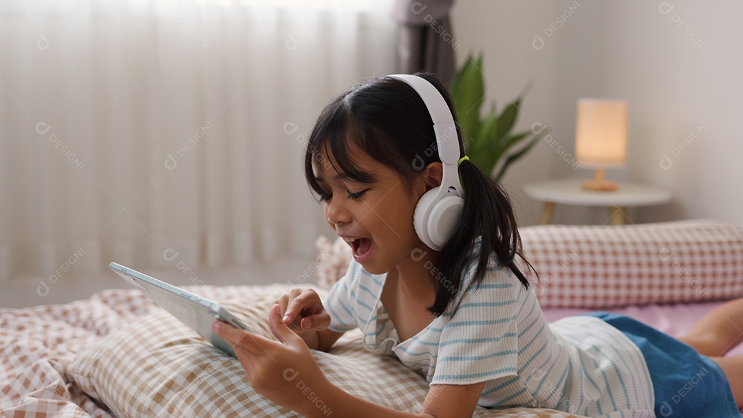 Garota asiática feliz deitada na cama usando tablet com fone de ouvido