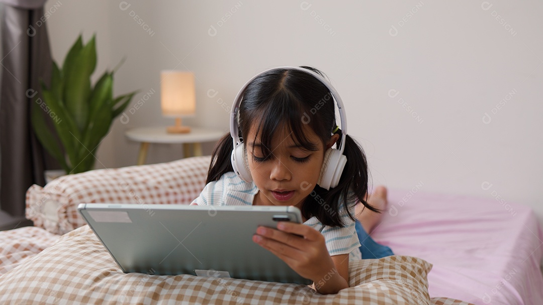 Garota asiática feliz deitada na cama usando tablet com fone de ouvido