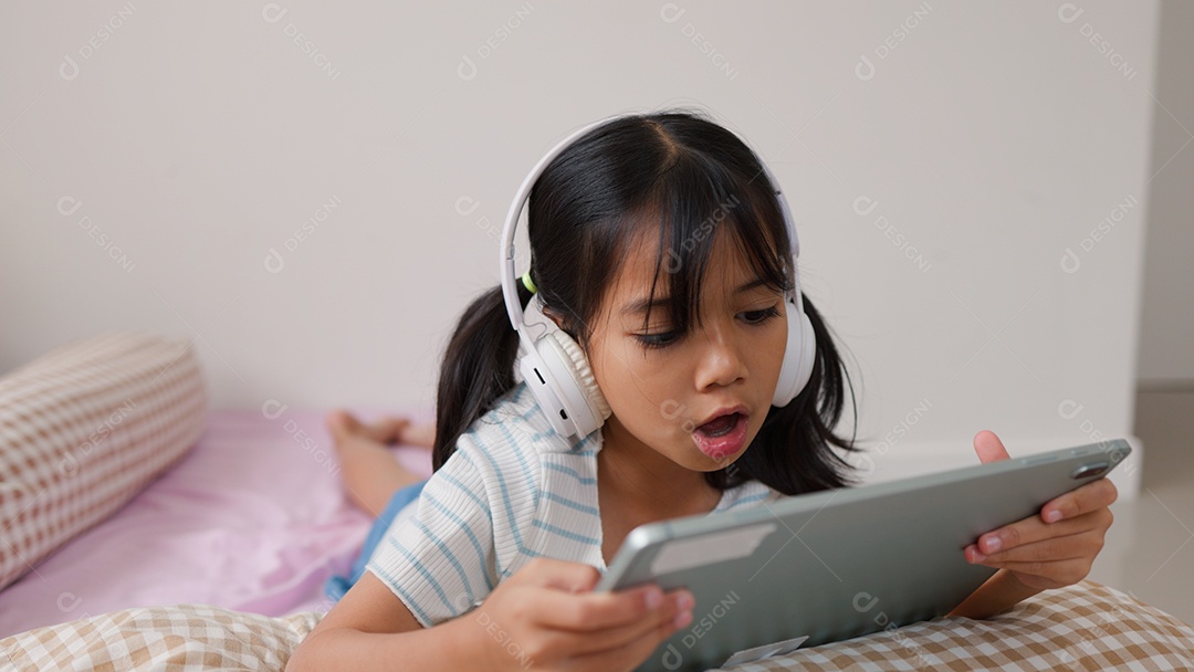 Garota asiática feliz deitada na cama usando tablet com fone de ouvido