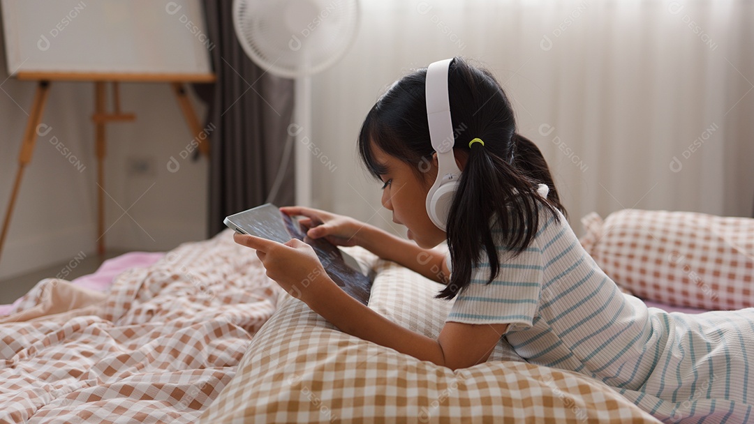 Garota asiática feliz deitada na cama usando tablet com fone de ouvido