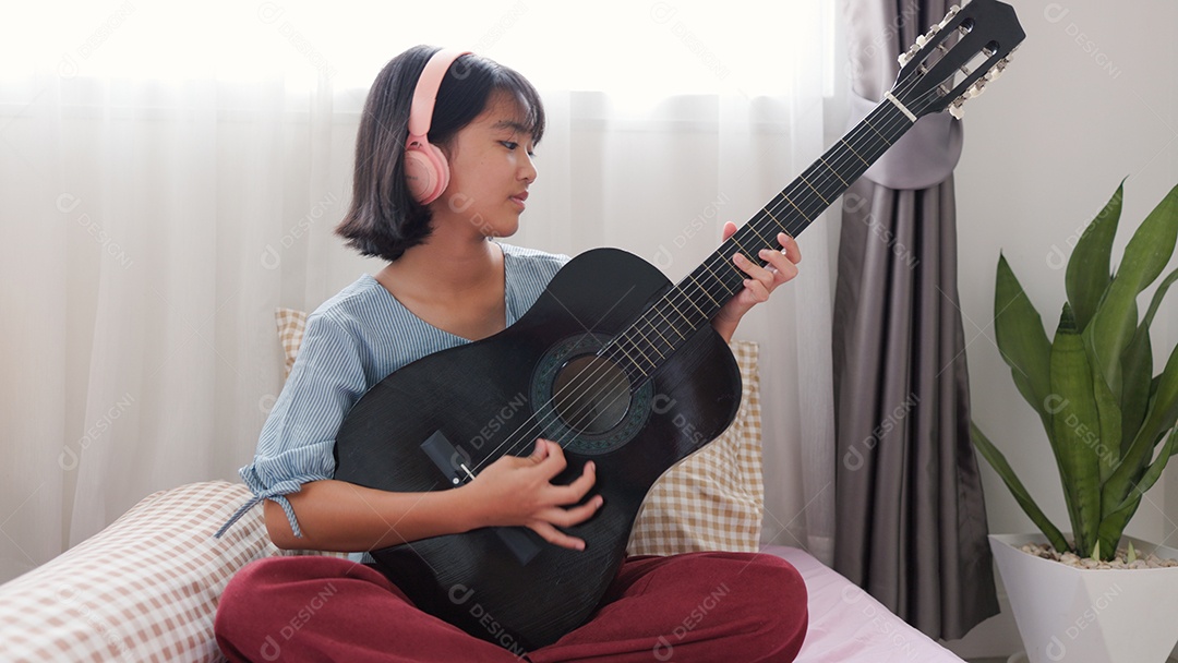 Menina asiática sentada na cama tocando violão com fones de ouvido