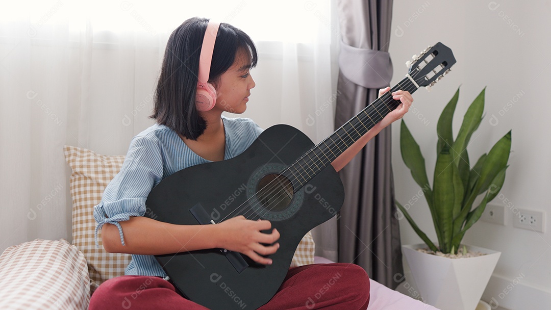 Menina asiática sentada na cama tocando violão com fones de ouvido