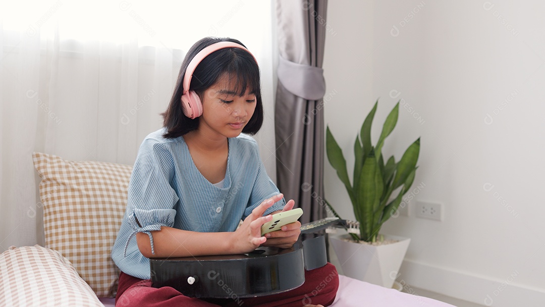 Menina asiática sentada na cama mechendo em smartphone com fones de ouvido