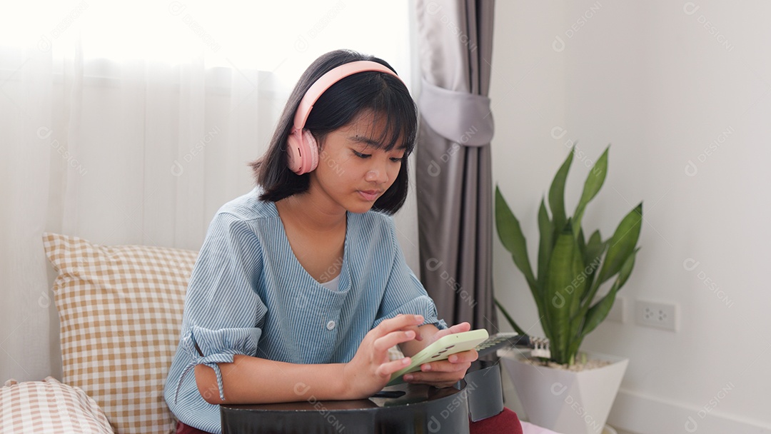 Menina asiática sentada na cama mechendo em smartphone com fones de ouvido
