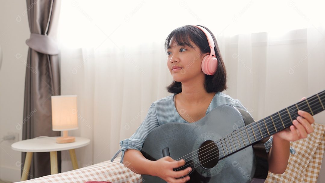 Menina asiática sentada na cama tocando violão com fones de ouvido