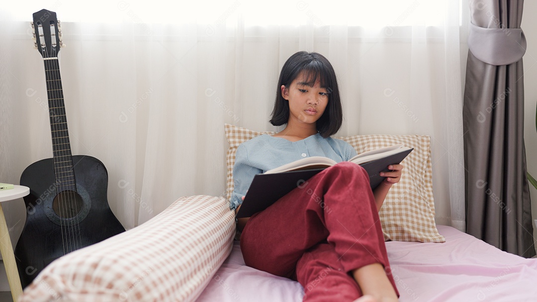 Menina asiática lendo um livro enquanto está sentada na cama em um quarto aconchegante