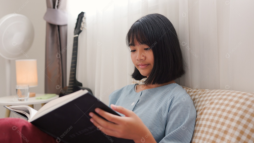 Menina asiática lendo um livro enquanto está sentada na cama em um quarto aconchegante