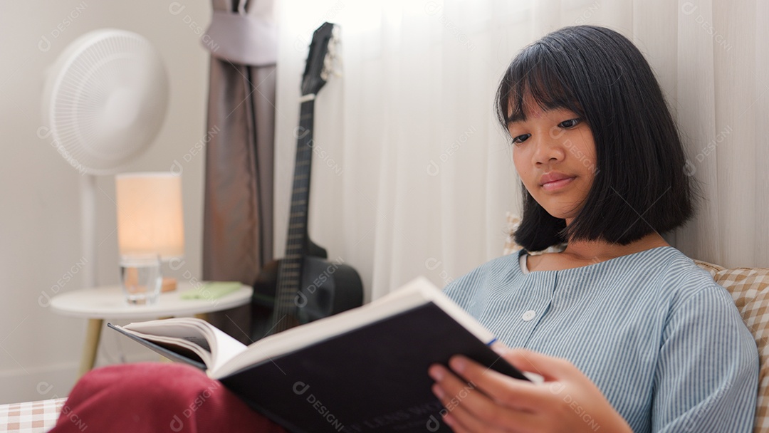 Menina asiática lendo um livro enquanto está sentada na cama em um quarto aconchegante