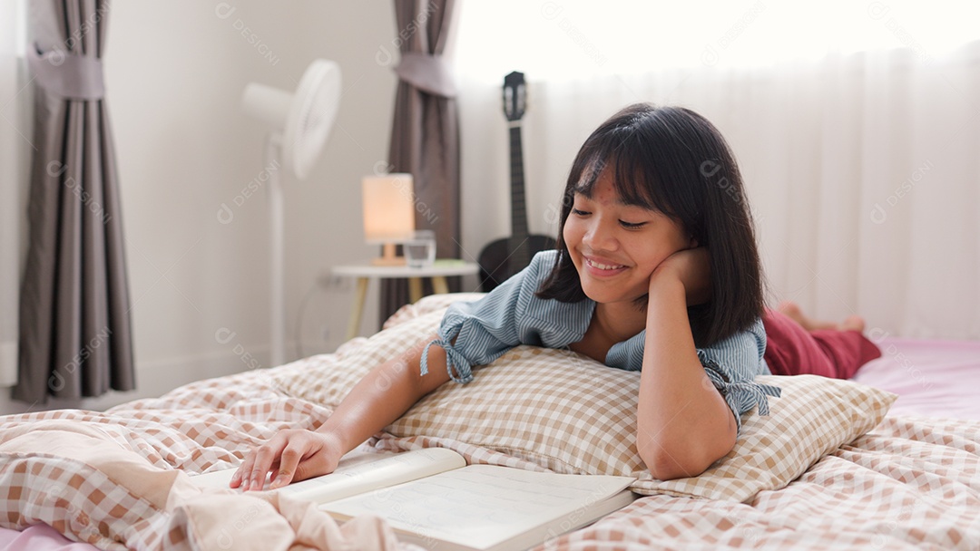 Menina asiática lendo um livro enquanto está sentada na cama em um quarto aconchegante