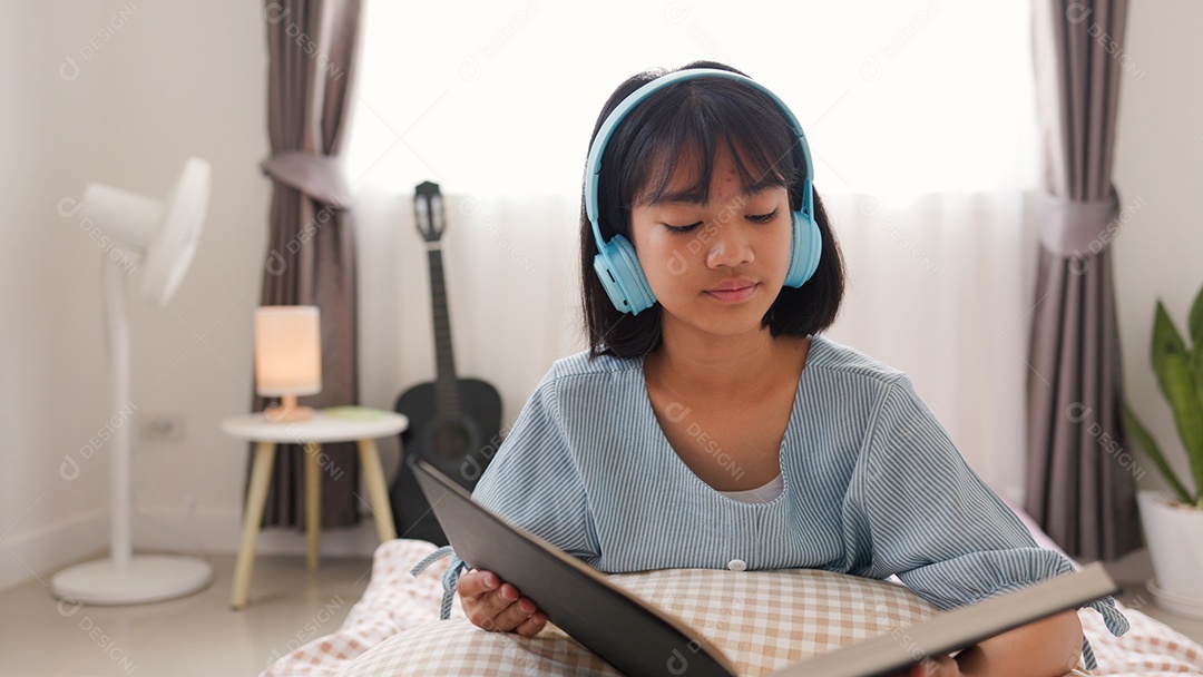 Menina asiática lendo um livro enquanto está sentada na cama em um quarto aconchegante