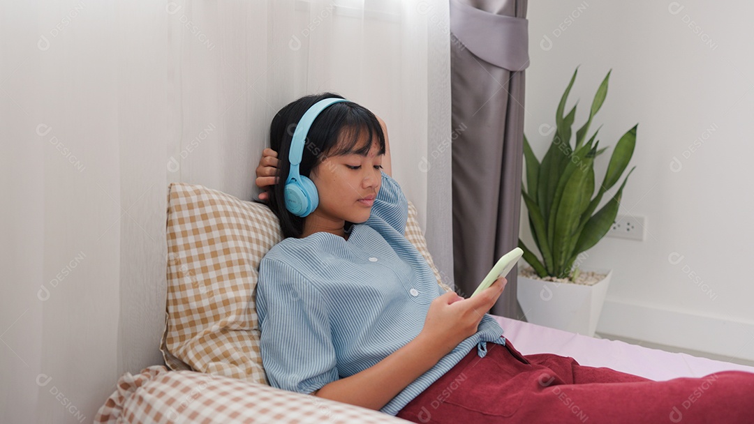 Menina asiática sentada na cama mechendo em smartphone com fones de ouvido