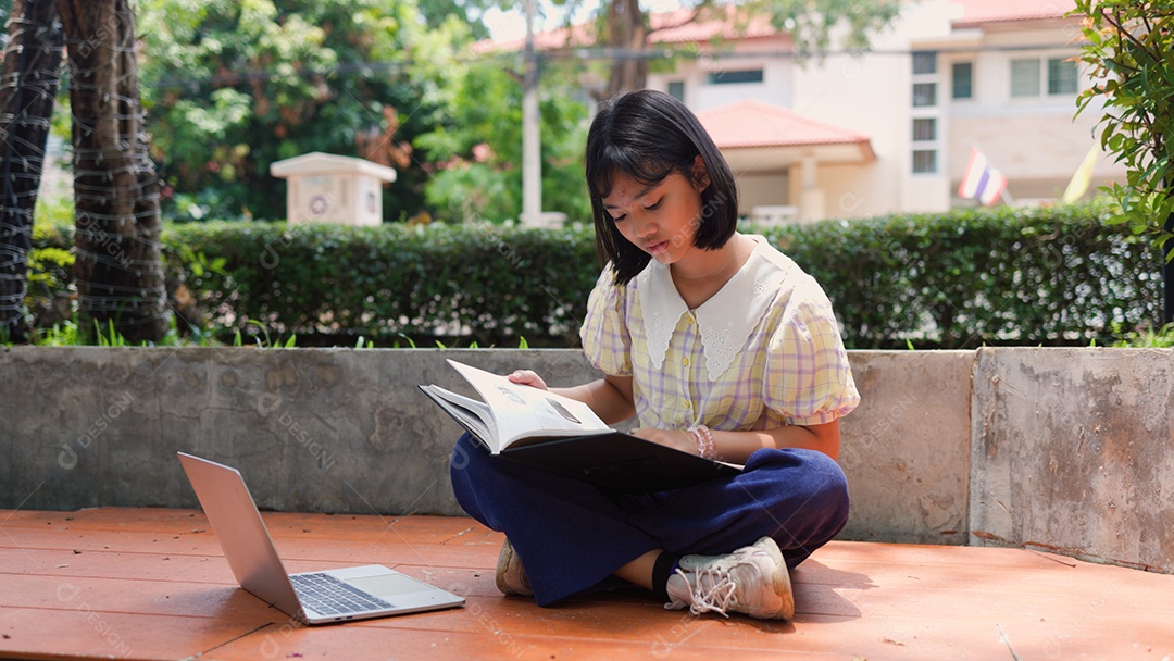 Adolescente asiática estudando ao ar livre usando um laptop e um livro