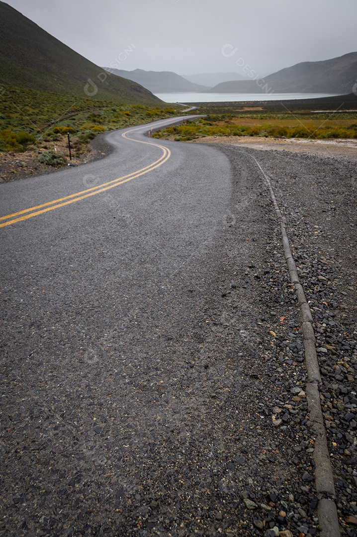 A imagem mostra uma estrada asfaltada e sinuosa que se estende por um vale montanhoso, com um lago ou rio visível ao fundo