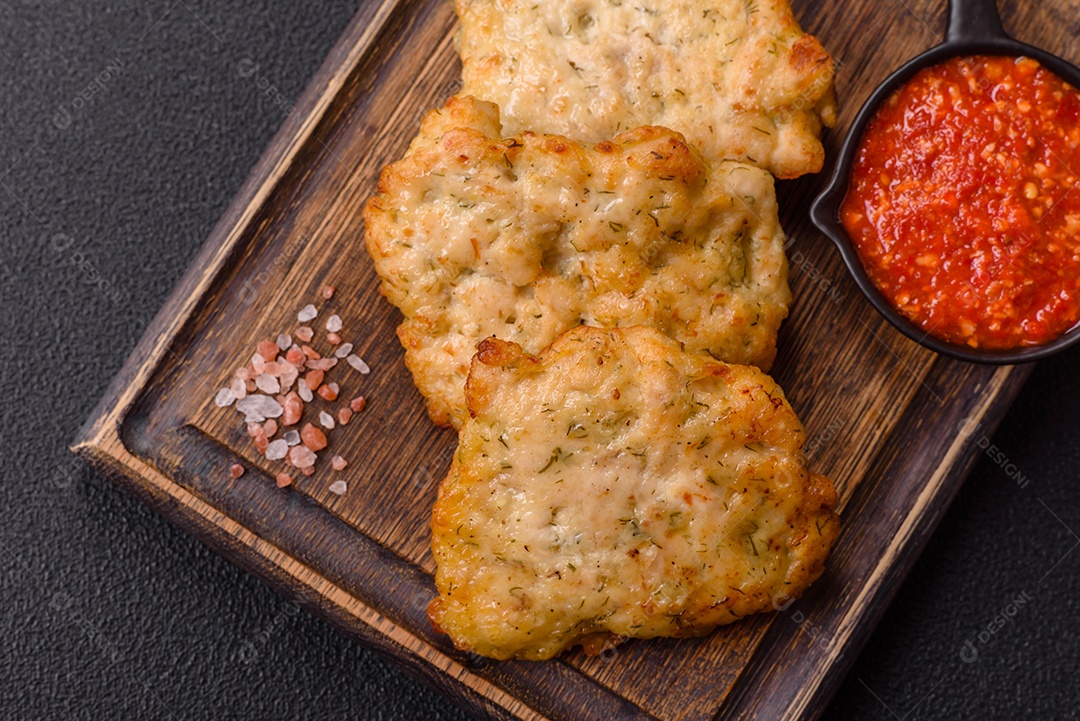 A imagem mostra pedaços de frango frito servidos em uma tábua de madeira com temperos e molho.