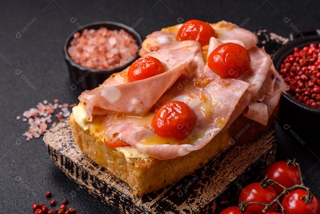 A imagem mostra um sanduíche de brioche com presunto, queijo derretido e tomates cereja, polvilhado com sal e pimenta.