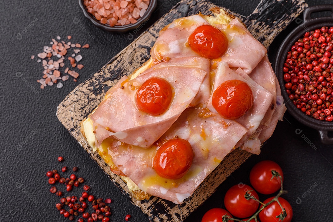 A imagem mostra um sanduíche de brioche com presunto, queijo derretido e tomates cereja, polvilhado com sal e pimenta.
