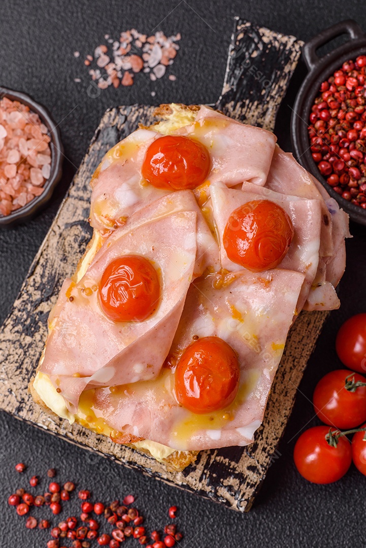 A imagem mostra um sanduíche de brioche com presunto, queijo derretido e tomates cereja, polvilhado com sal e pimenta.
