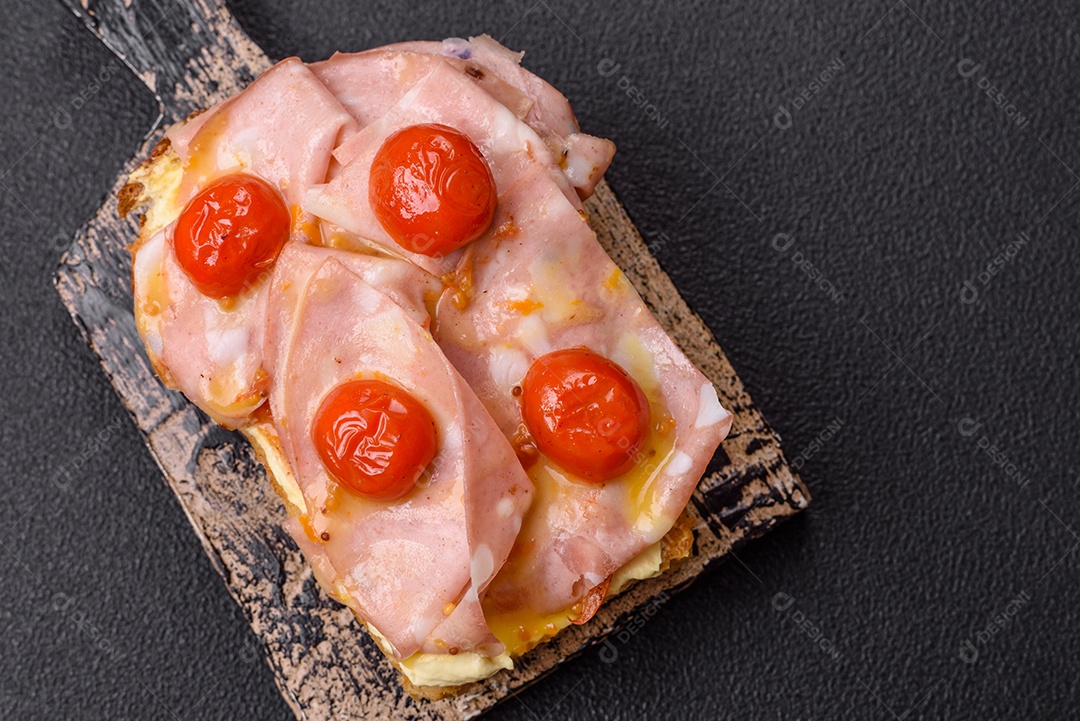 A imagem mostra um sanduíche de brioche com presunto, queijo derretido e tomates cereja, polvilhado com sal e pimenta.