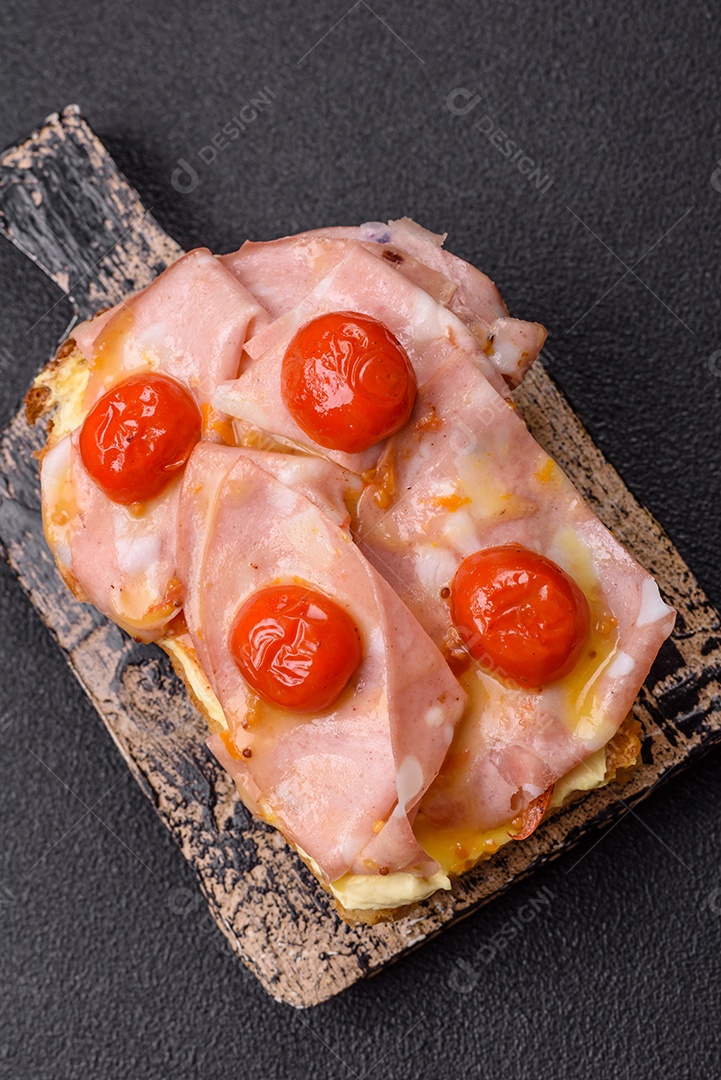 A imagem mostra um sanduíche de brioche com presunto, queijo derretido e tomates cereja, polvilhado com sal e pimenta.