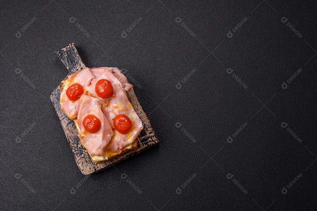 A imagem mostra um sanduíche de brioche com presunto, queijo derretido e tomates cereja, polvilhado com sal e pimenta.