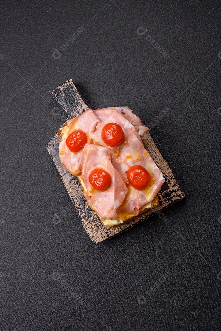 A imagem mostra um sanduíche de brioche com presunto, queijo derretido e tomates cereja, polvilhado com sal e pimenta.