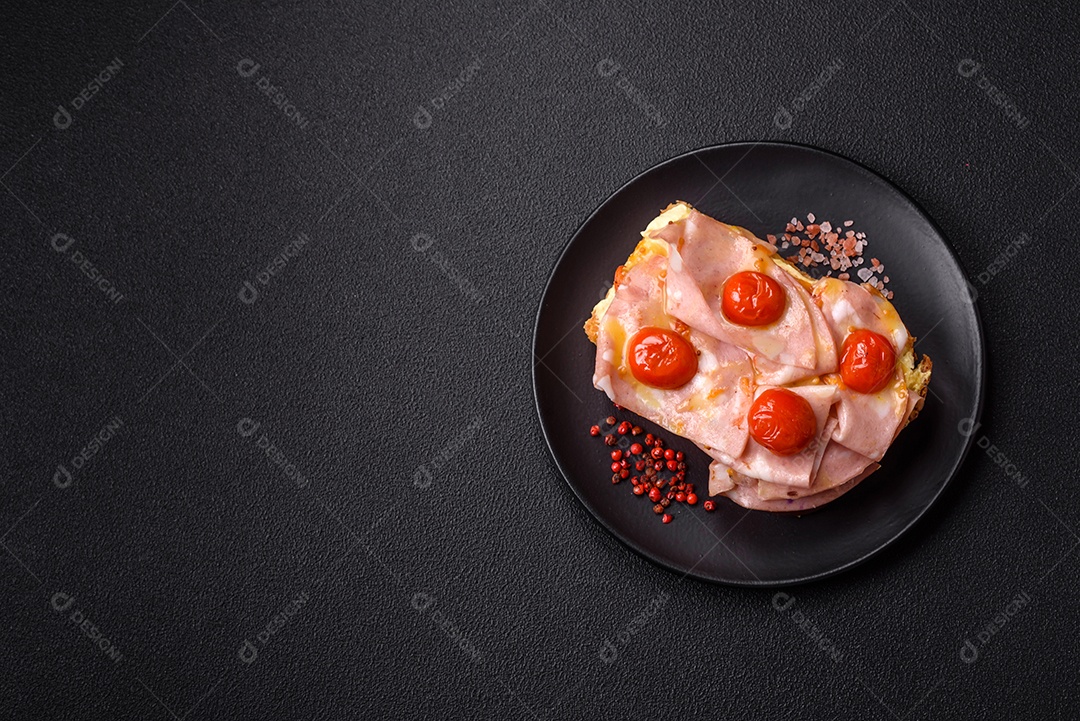 A imagem mostra um sanduíche de brioche com presunto, queijo derretido e tomates cereja, polvilhado com sal e pimenta.