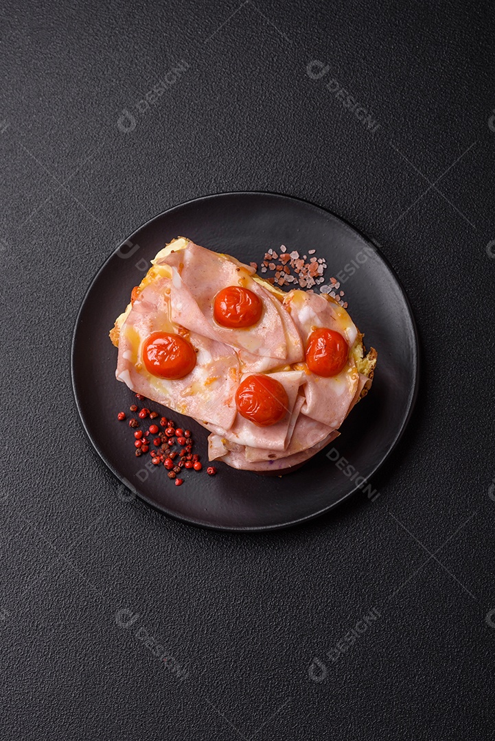 A imagem mostra um sanduíche de brioche com presunto, queijo derretido e tomates cereja, polvilhado com sal e pimenta.