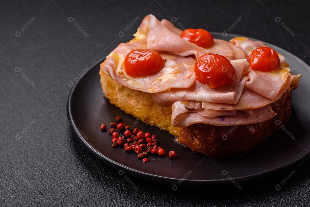 A imagem mostra um sanduíche de brioche com presunto, queijo derretido e tomates cereja, polvilhado com sal e pimenta.