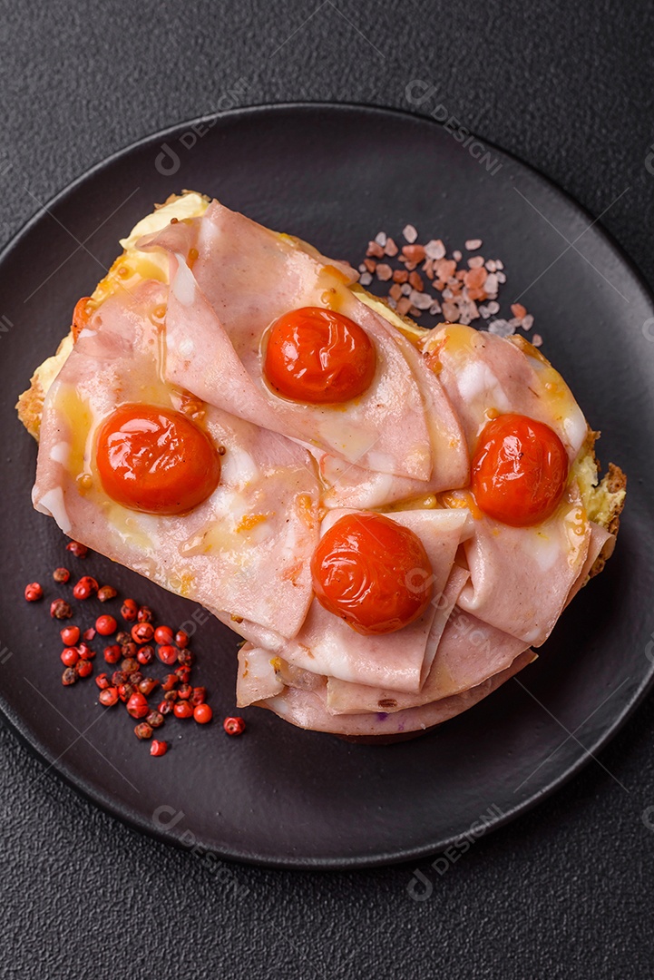 A imagem mostra um sanduíche de brioche com presunto, queijo derretido e tomates cereja, polvilhado com sal e pimenta.