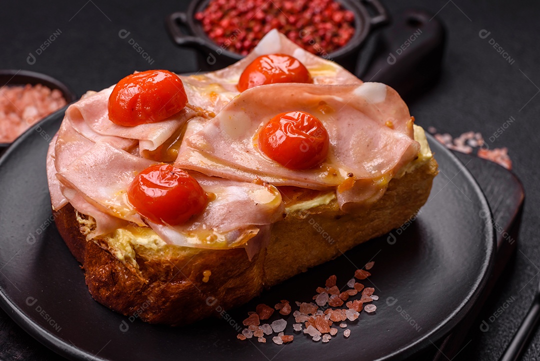 A imagem mostra um sanduíche de brioche com presunto, queijo derretido e tomates cereja, polvilhado com sal e pimenta.