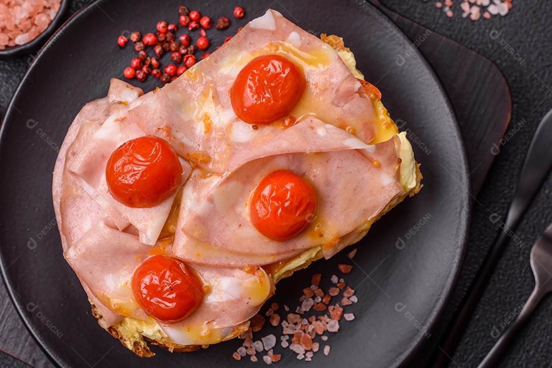 A imagem mostra um sanduíche de brioche com presunto, queijo derretido e tomates cereja, polvilhado com sal e pimenta.