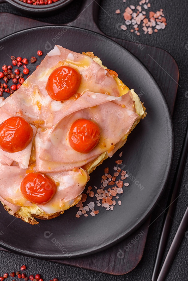 A imagem mostra um sanduíche de brioche com presunto, queijo derretido e tomates cereja, polvilhado com sal e pimenta.