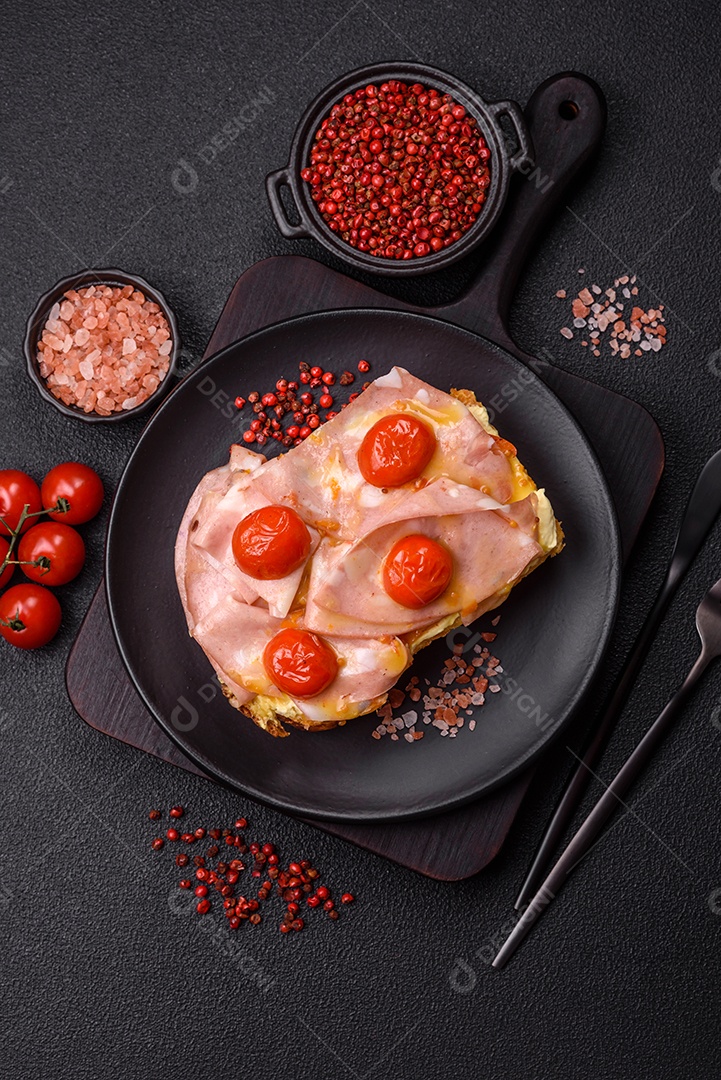 A imagem mostra um sanduíche de brioche com presunto, queijo derretido e tomates cereja, polvilhado com sal e pimenta.