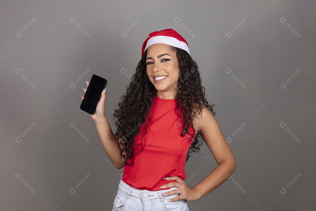 Linda jovem morena cabelo cacheado usando gorro do papai noel segurando celular smartphone