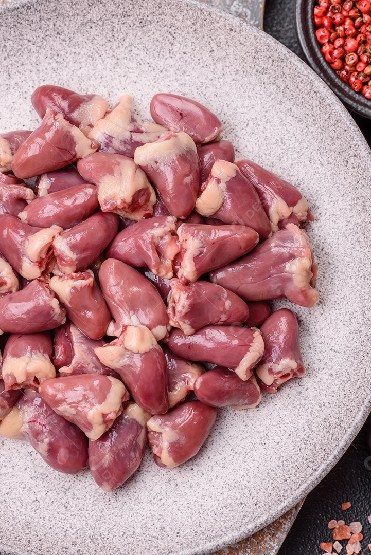 A imagem mostra corações de frango crus em uma tábua de madeira, acompanhados de pimenta rosa e tomates-cereja.