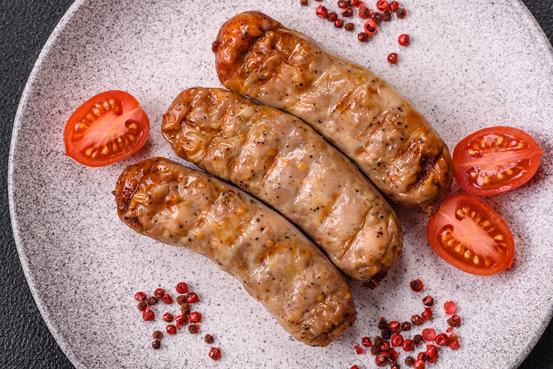 Três linguiças ou salsichas grelhadas são apresentadas em um prato, com temperos e tomates-cereja cortados.
